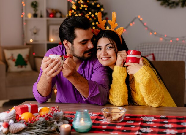 Giovane e bella coppia uomo e donna seduti al tavolo con tazze di tè felici nell'amore nella stanza decorata di natale con l'albero di natale in background