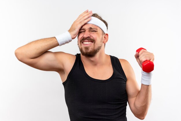 Giovane e bello sportivo dolorante che indossa fascia e braccialetti che tengono il manubrio e si mettono la mano sulla testa isolata sul muro bianco