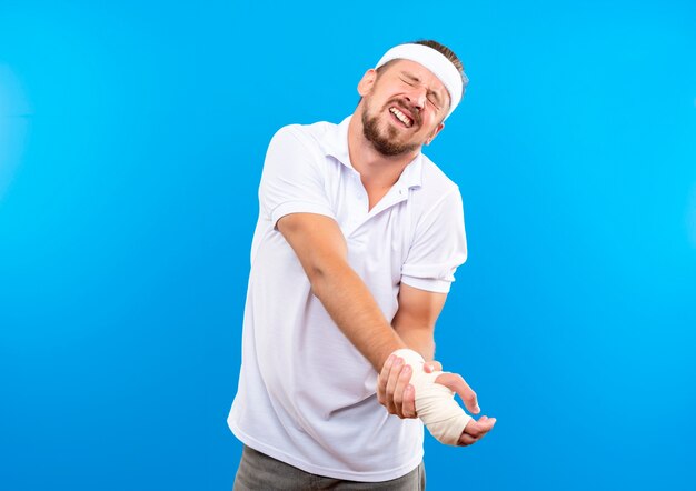 Giovane e bello sportivo dolorante che indossa fascia e braccialetti che tengono il polso ferito avvolto con una benda con gli occhi chiusi isolato sulla parete blu con spazio di copia