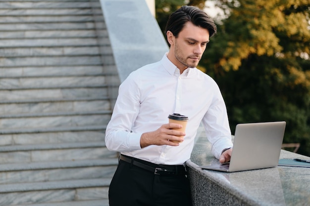 Giovane e bello uomo bruna barbuto serio in camicia bianca che tiene una tazza di caffè per andare in mano a lavorare premurosamente sul computer portatile nel parco cittadino da solo