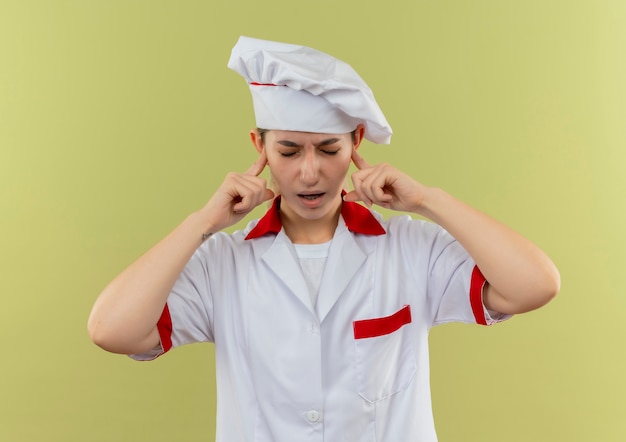 Giovane e graziosa cuoca infastidita in uniforme da chef che mette le dita nelle orecchie con gli occhi chiusi isolati sul muro verde