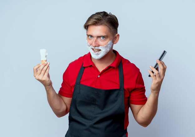 giovane eccitato barbiere maschio biondo in uniforme spalmato di viso con schiuma da barba tenendo il pennello da barba