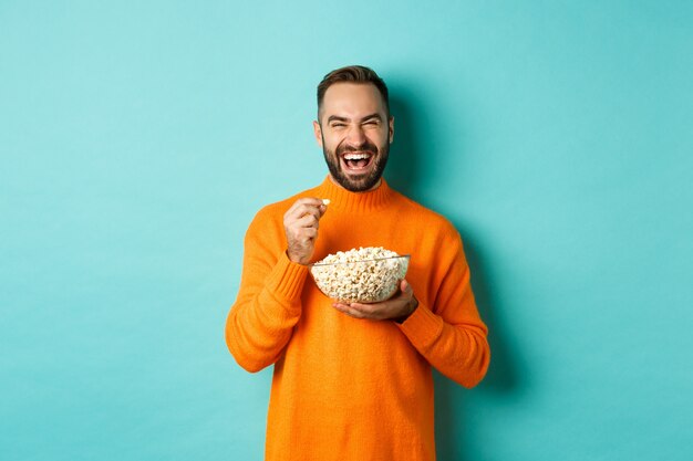 Giovane eccitato che guarda film interessante sullo schermo televisivo, mangia popcorn e sembra stupito