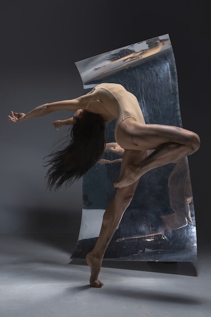 Giovane ed elegante ballerina moderna sulla parete grigia con i riflessi di specchio e illusione sulla superficie