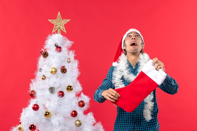 Giovane emotivo con il cappello di Babbo Natale in una camicia spogliata blu e tenendo il calzino di Natale guardando sopra