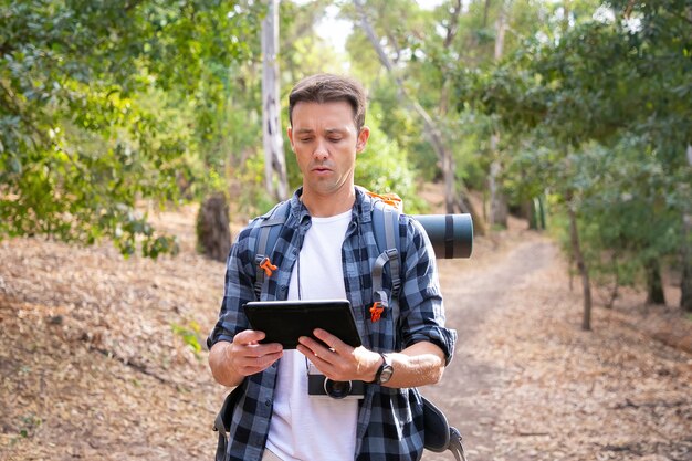 Giovane escursionista zaino in spalla, tenendo tablet e guardando la mappa. Viaggiatore attraente caucasico che cammina sulla strada nel bosco. Concetto di turismo, avventura e vacanze estive con lo zaino in spalla