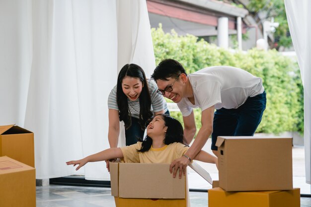 Giovane famiglia asiatica felice divertendosi ridendo entrando nella nuova casa. Genitori giapponesi madre e padre che sorridono aiutando la guida emozionante della bambina che si siede in scatola di cartone. Nuova proprietà e trasferimento.