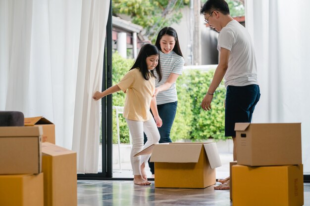 Giovane famiglia asiatica felice divertendosi ridendo entrando nella nuova casa. Genitori giapponesi madre e padre che sorridono aiutando la guida emozionante della bambina che si siede in scatola di cartone. Nuova proprietà e trasferimento.