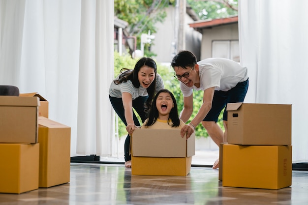 Giovane famiglia asiatica felice divertendosi ridendo entrando nella nuova casa. Genitori giapponesi madre e padre che sorridono aiutando la guida emozionante della bambina che si siede in scatola di cartone. Nuova proprietà e trasferimento.