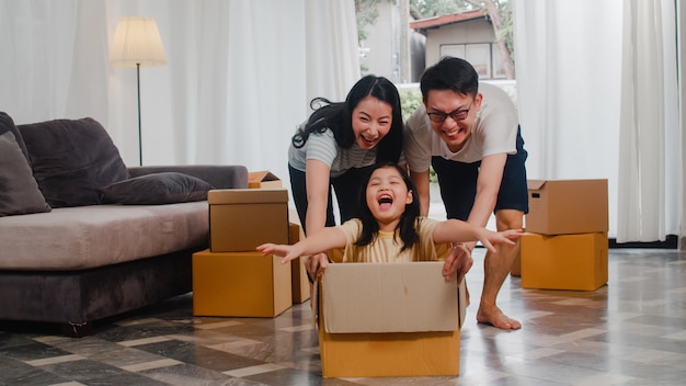 Giovane famiglia asiatica felice divertendosi ridendo entrando nella nuova casa. Genitori giapponesi madre e padre che sorridono aiutando la guida emozionante della bambina che si siede in scatola di cartone. Nuova proprietà e trasferimento.