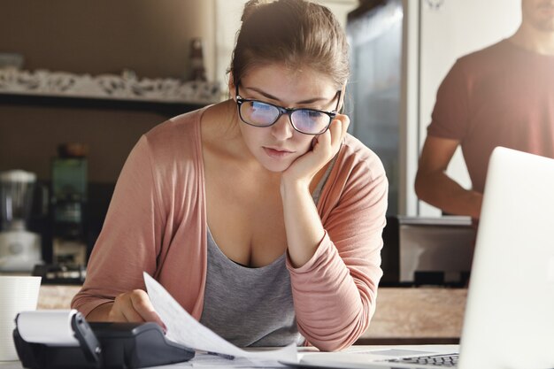 Giovane famiglia caucasica che affronta il problema del credito. Moglie stressata che tiene pezzo di carta e calcola le finanze, facendo del suo meglio per tagliare le spese domestiche usando la calcolatrice e il computer portatile generico