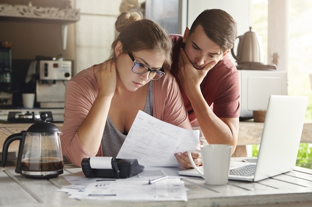 Giovane famiglia caucasica con problemi di debito, non in grado di pagare il proprio prestito. La femmina in vetri e l'uomo castana che studiano la forma di carta incassano mentre gestiscono insieme il bilancio domestico nell'interno della cucina