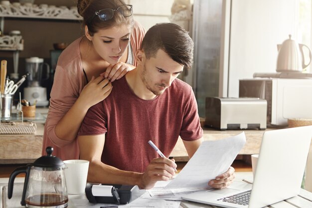 Giovane famiglia infelice che paga le bollette online sul computer portatile. Stressato uomo seduto al tavolo con documenti e calcolatrice, compilando documenti, calcolando le spese domestiche insieme alla sua bella moglie