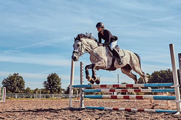 Giovane fantino femmina sul cavallo grigio dapple che salta sopra l'ostacolo nell'arena aperta.