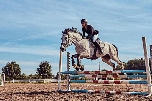 Giovane fantino femmina sul cavallo grigio dapple che salta sopra l'ostacolo nell'arena aperta.