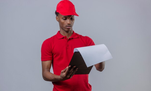 Giovane fattorino afroamericano che indossa la camicia e il cappuccio di polo rossi che esaminano lavagna per appunti con il fronte serio sopra bianco isolato