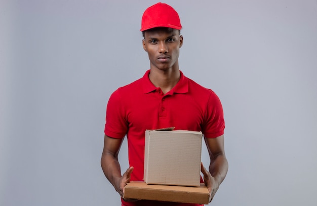 Giovane fattorino afroamericano che indossa la pila della tenuta della camicia e del cappuccio di polo rosso di scatole di cartone che esaminano macchina fotografica con il fronte serio sopra bianco isolato