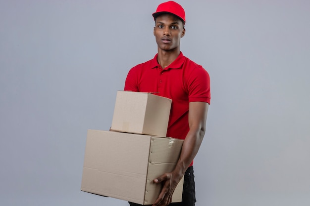 Giovane fattorino afroamericano che indossa la pila della tenuta della camicia e del cappuccio di polo rosso di scatole di cartone che esaminano macchina fotografica con il fronte serio sopra bianco isolato