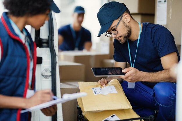 Giovane fattorino che utilizza la tavoletta digitale per la scansione del codice a barre sull'etichetta del pacco mentre si prepara per la spedizione con il suo collega