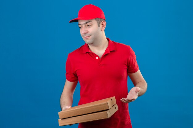 Giovane fattorino in scatole della pizza della tenuta della camicia e del cappuccio di polo rosso che guardano da parte con il fronte indagatore sopra la parete blu isolata