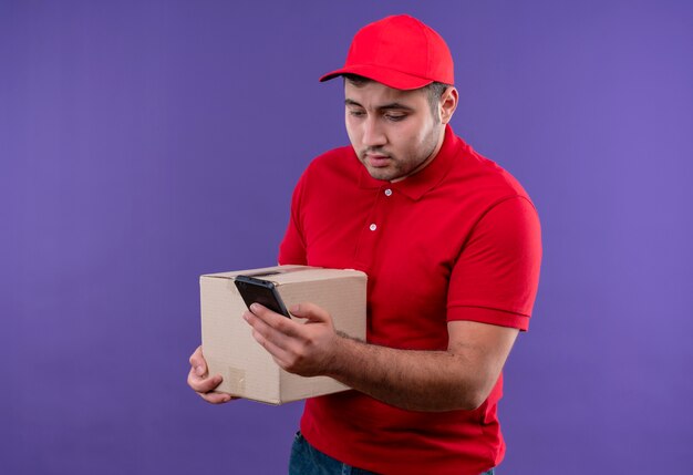 Giovane fattorino in uniforme rossa e berretto che tiene il pacchetto della scatola guardando lo schermo del suo smartphone che sembra confuso in piedi sopra la parete viola