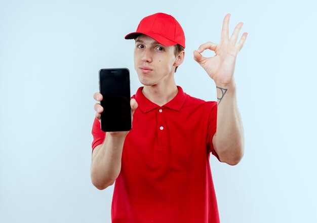 Giovane fattorino in uniforme rossa e cappuccio che mostra smartphone che mostra segno giusto in piedi sopra il muro bianco