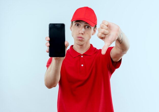 Giovane fattorino in uniforme rossa e cappuccio che mostra smartphone guardando in avanti dispiaciuto mostrando i pollici verso il basso in piedi sopra il muro bianco