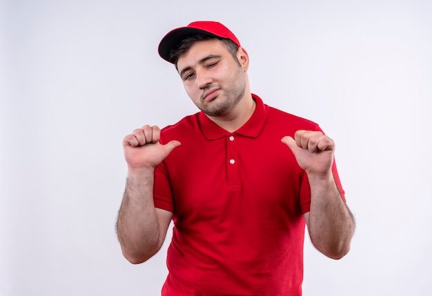 Giovane fattorino in uniforme rossa e cappuccio che punta con i pollici a se stesso, soddisfatto di sé e orgoglioso in piedi sopra il muro bianco