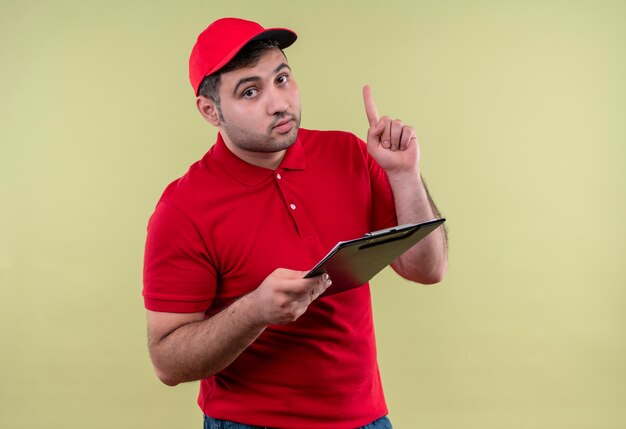 Giovane fattorino in uniforme rossa e cappuccio che tiene appunti rivolto verso l'alto con il dito guardando fiducioso in piedi sopra la parete verde