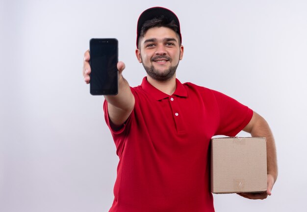 Giovane fattorino in uniforme rossa e cappuccio che tiene il pacchetto della scatola che mostra smartphone che guarda l'obbiettivo con un sorriso sicuro