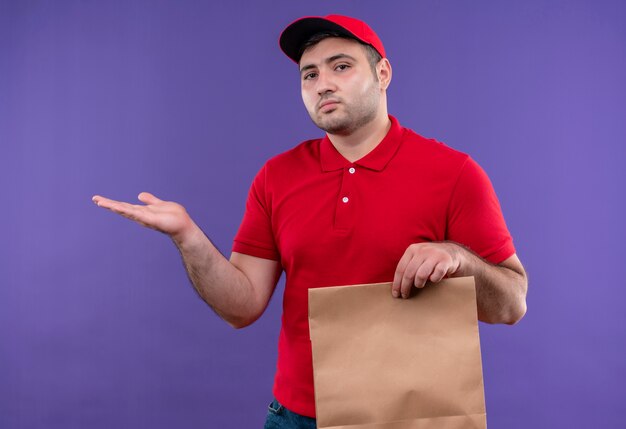 Giovane fattorino in uniforme rossa e cappuccio che tiene il pacchetto di carta che presenta lo spazio della copia con il braccio della mano che sta sopra la parete viola