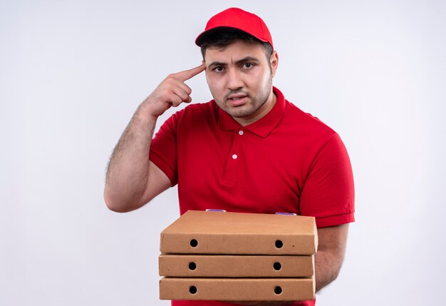 Giovane fattorino in uniforme rossa e cappuccio che tiene le scatole della pizza che indica con il dito il suo tempio che sembra fiducioso concentrato su un compito in piedi sopra il muro bianco