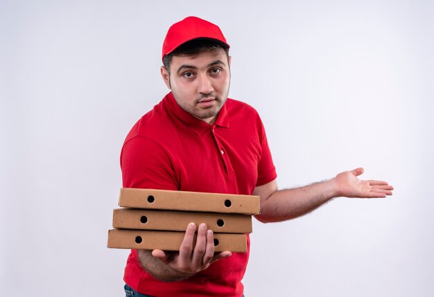 Giovane fattorino in uniforme rossa e cappuccio che tiene le scatole per pizza che presentano con il braccio della sua mano in piedi sul muro bianco