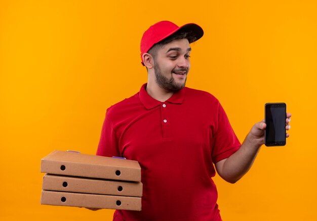 Giovane fattorino in uniforme rossa e cappuccio che tiene pila di scatole per pizza che mostra smartphone guardandolo con il sorriso sul viso