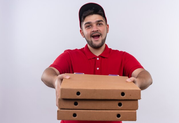 Giovane fattorino in uniforme rossa e cappuccio che tiene pila di scatole per pizza guardando fiducioso in generale
