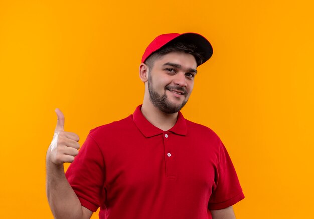 Giovane fattorino in uniforme rossa e cappuccio sorridente amichevole che mostra i pollici in su