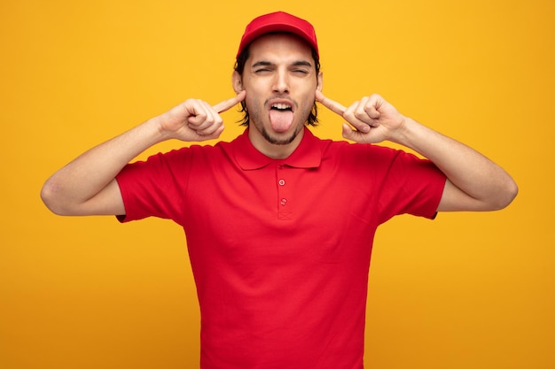 giovane fattorino infastidito che indossa uniforme e cappuccio che mette le dita nelle orecchie guardando la fotocamera che mostra la lingua isolata su sfondo giallo