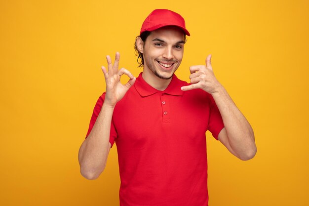 giovane fattorino sorridente che indossa uniforme e cappuccio guardando la fotocamera che mostra il gesto di chiamata e il segno ok isolato su sfondo giallo