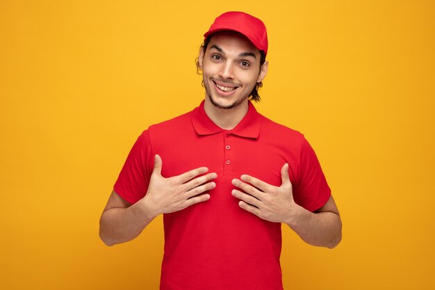 giovane fattorino sorridente che indossa uniforme e cappuccio guardando la fotocamera che punta su se stesso con le mani isolate su sfondo giallo