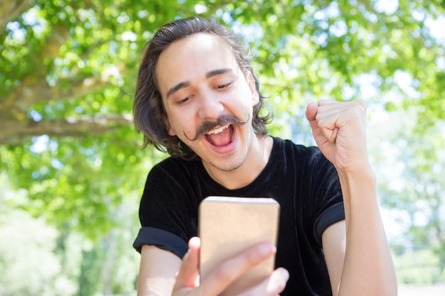 Giovane felice che esamina smartphone in parco.