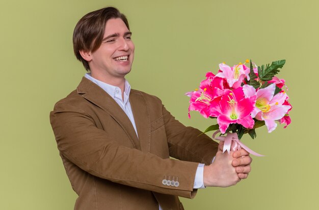 Giovane felice pazzo che tiene il mazzo di fiori che osserva da parte sorridendo allegramente andando a congratularsi con la giornata internazionale della donna in piedi sopra la parete verde