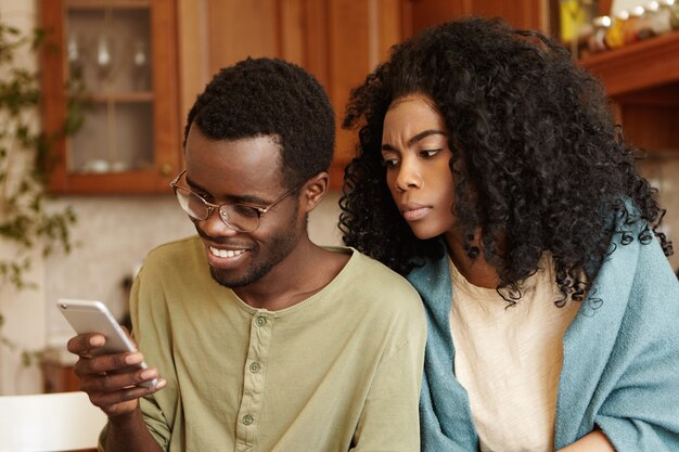 Giovane femmina afroamericana possessiva ossessionata che osserva sopra la spalla di suo marito, provando a leggere i messaggi sul suo telefono cellulare. Persone, relazioni, privacy, infedeltà e tecnologie moderne