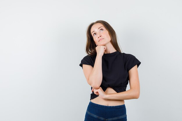 Giovane femmina appoggiando il mento sul pugno in camicetta nera, pantaloni e guardando speranzoso.