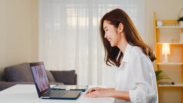 Giovane femmina asiatica di affari che per mezzo della videochiamata del computer portatile che parla con il papà e la mamma della famiglia mentre lavora da casa al salone. Autoisolamento, allontanamento sociale, quarantena per la prevenzione del coronavirus.