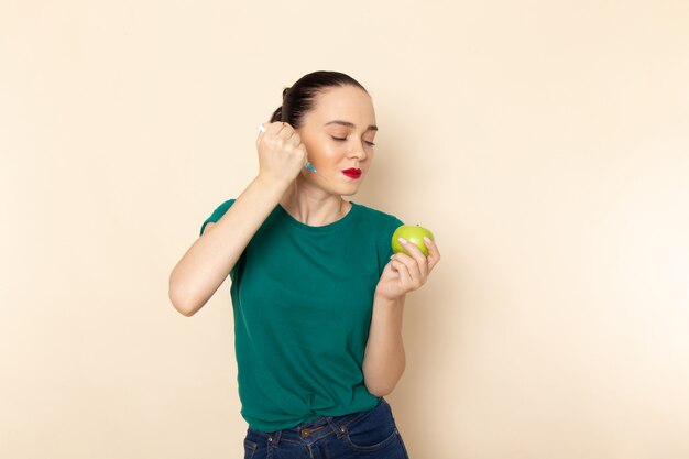 Giovane femmina attraente di vista frontale in camicia verde scuro e blue jeans che iniettano mela sul beige