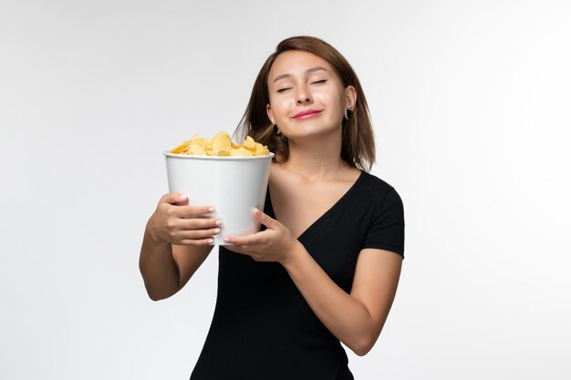 Giovane femmina attraente di vista frontale in canestro nero della tenuta della camicia con le patatine fritte sulla superficie bianca