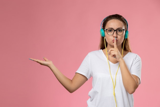 Giovane femmina attraente di vista frontale in maglietta bianca che ascolta la musica che posa mostrando il segno di silenzio sui precedenti rosa