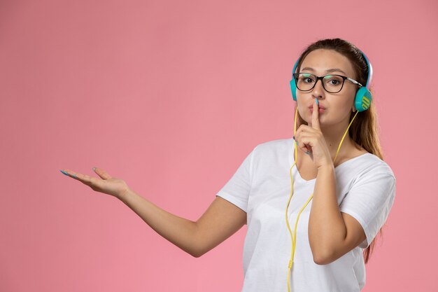 Giovane femmina attraente di vista frontale in maglietta bianca che ascolta la musica con gli auricolari che mostrano il segno di silenzio sui precedenti rosa