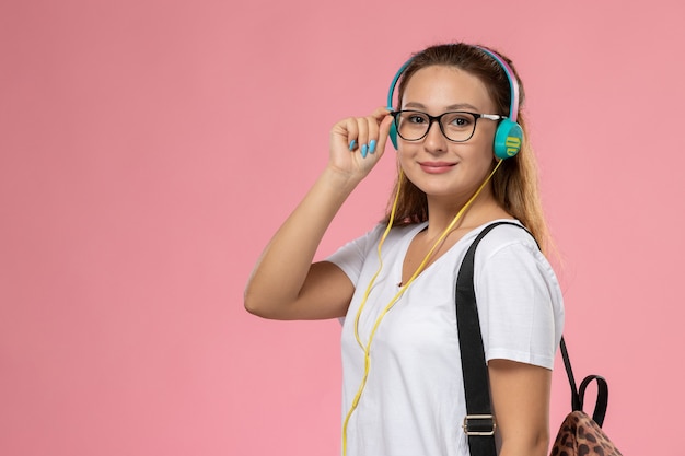 Giovane femmina attraente di vista frontale in maglietta bianca che ascolta la musica con gli auricolari che sorridono sullo scrittorio rosa