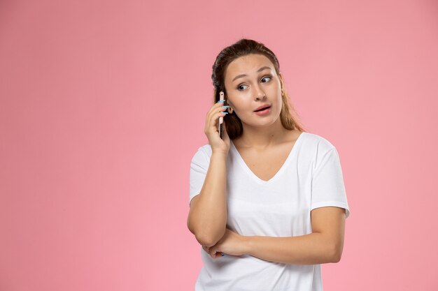 Giovane femmina attraente di vista frontale in maglietta bianca che parla sul telefono sui precedenti rosa